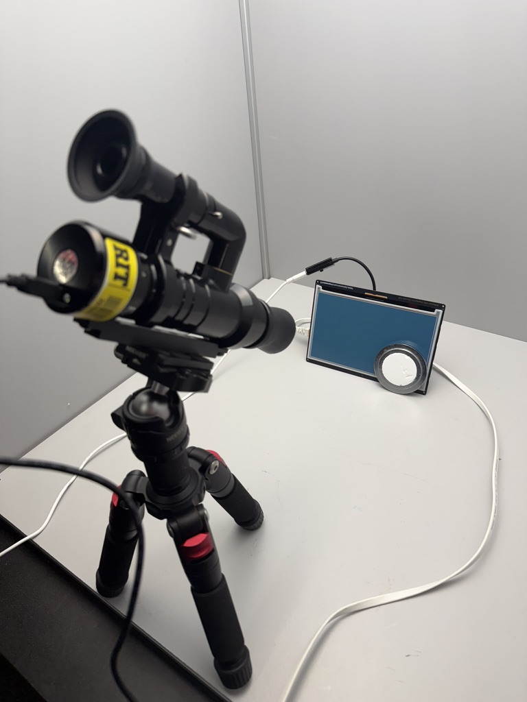 Colorimetry Research CR-250 on a tripod facing the externally illuminated display, with a separate reference disk nearby